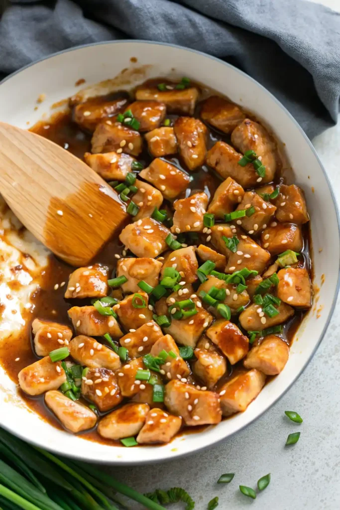 Teriyaki chicken cooking in a white skillet with wooden spatula, topped with sesame seeds and chopped green onions.
