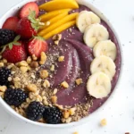 Thick acai bowl topped with banana slices, strawberries, mango strips, blackberries, and crunchy granola in a white bowl
