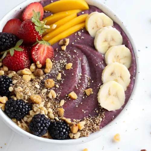Thick acai bowl topped with banana slices, strawberries, mango strips, blackberries, and crunchy granola in a white bowl