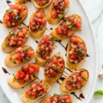 Toasted baguette slices topped with fresh tomato bruschetta and balsamic glaze on a white serving platter