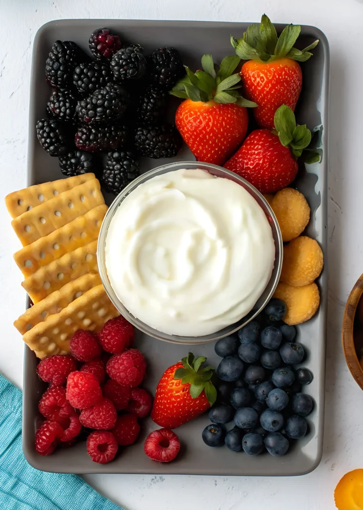 Easy fruit dip in a glass bowl served with strawberries, blackberries, blueberries, raspberries, crackers, and vanilla wafers on a gray platter.