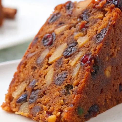 Close-up of a slice of moist fruit cake with visible dried fruits and nuts on a white plate
