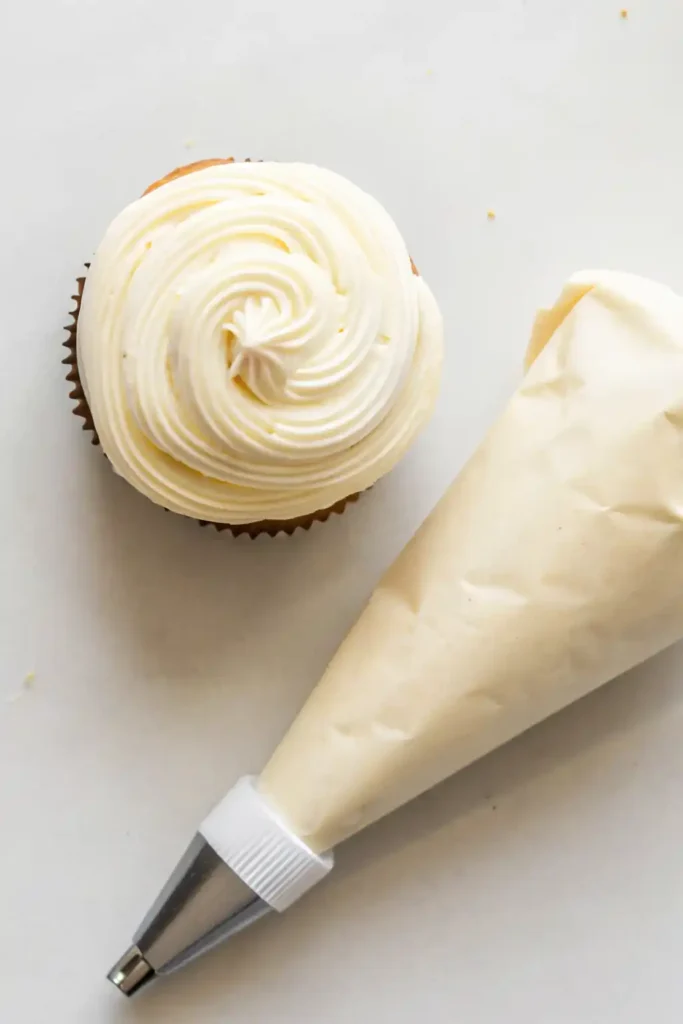 Vanilla cupcake with piped buttercream frosting next to a filled piping bag on a white surface