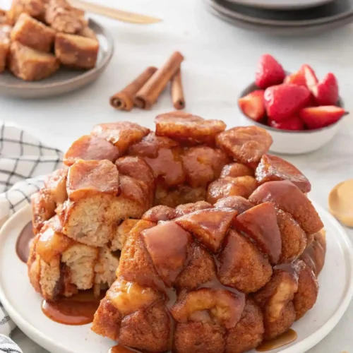 Homemade monkey bread on a white plate, glazed with caramel and surrounded by fresh strawberries, cinnamon sticks, and stacked dishes.