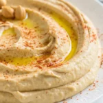 Smooth homemade hummus topped with olive oil, paprika, and chickpeas on a white plate