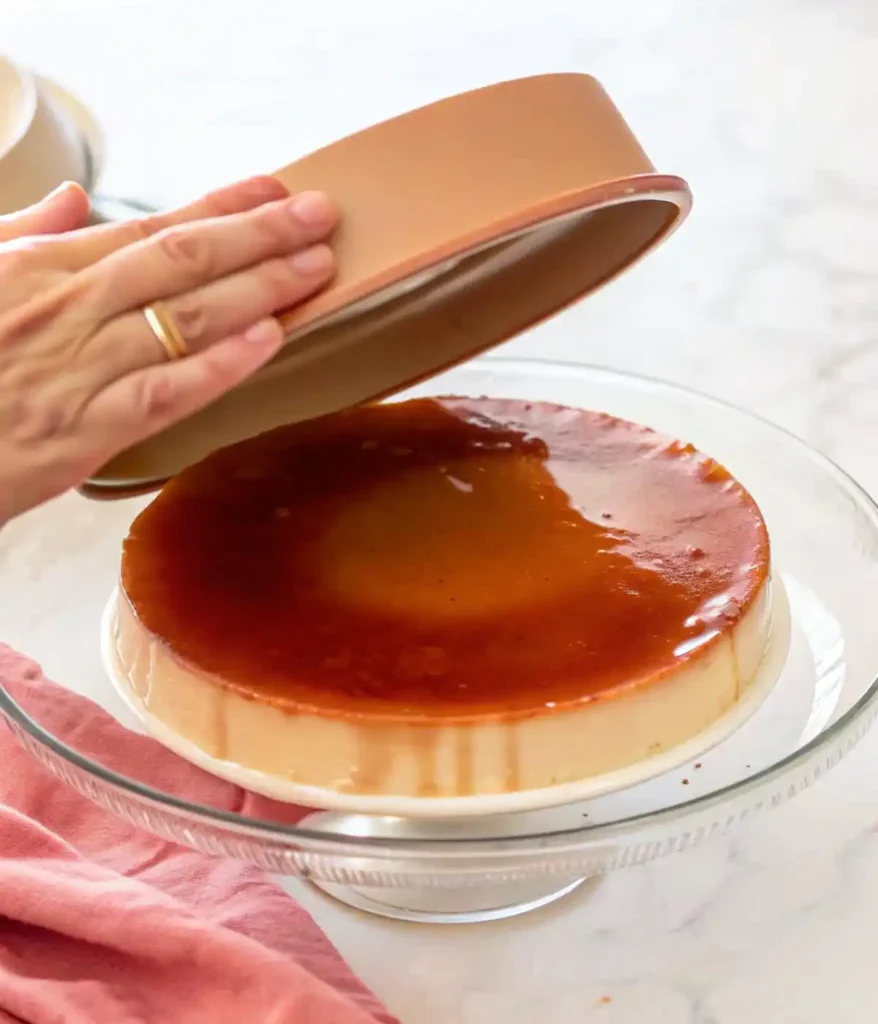 Freshly baked flan being released from a round pan onto a glass cake stand with caramel on top.