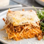 Slice of baked million dollar spaghetti with creamy cheese layers, meat sauce, and spaghetti noodles on a gray plate, served with a fresh green salad.