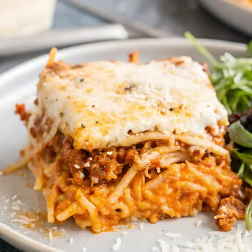 Slice of baked million dollar spaghetti with creamy cheese layers, meat sauce, and spaghetti noodles on a gray plate, served with a fresh green salad.