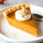 Slice of homemade pumpkin pie with whipped cream and cinnamon on top, served on a white plate.