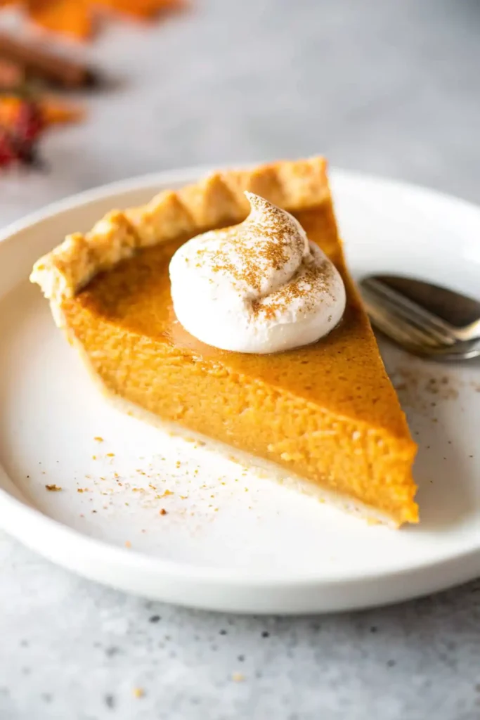 Slice of homemade pumpkin pie with whipped cream and cinnamon on top, served on a white plate.