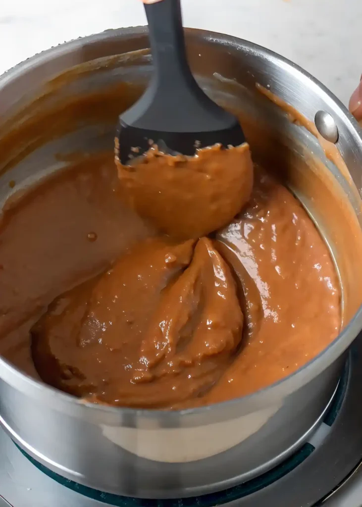 Thick brigadeiro mixture being stirred in a stainless steel saucepan using a black silicone spatula during cooking.