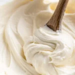 Smooth vanilla buttercream frosting being stirred with a wooden spoon in a glass bowl