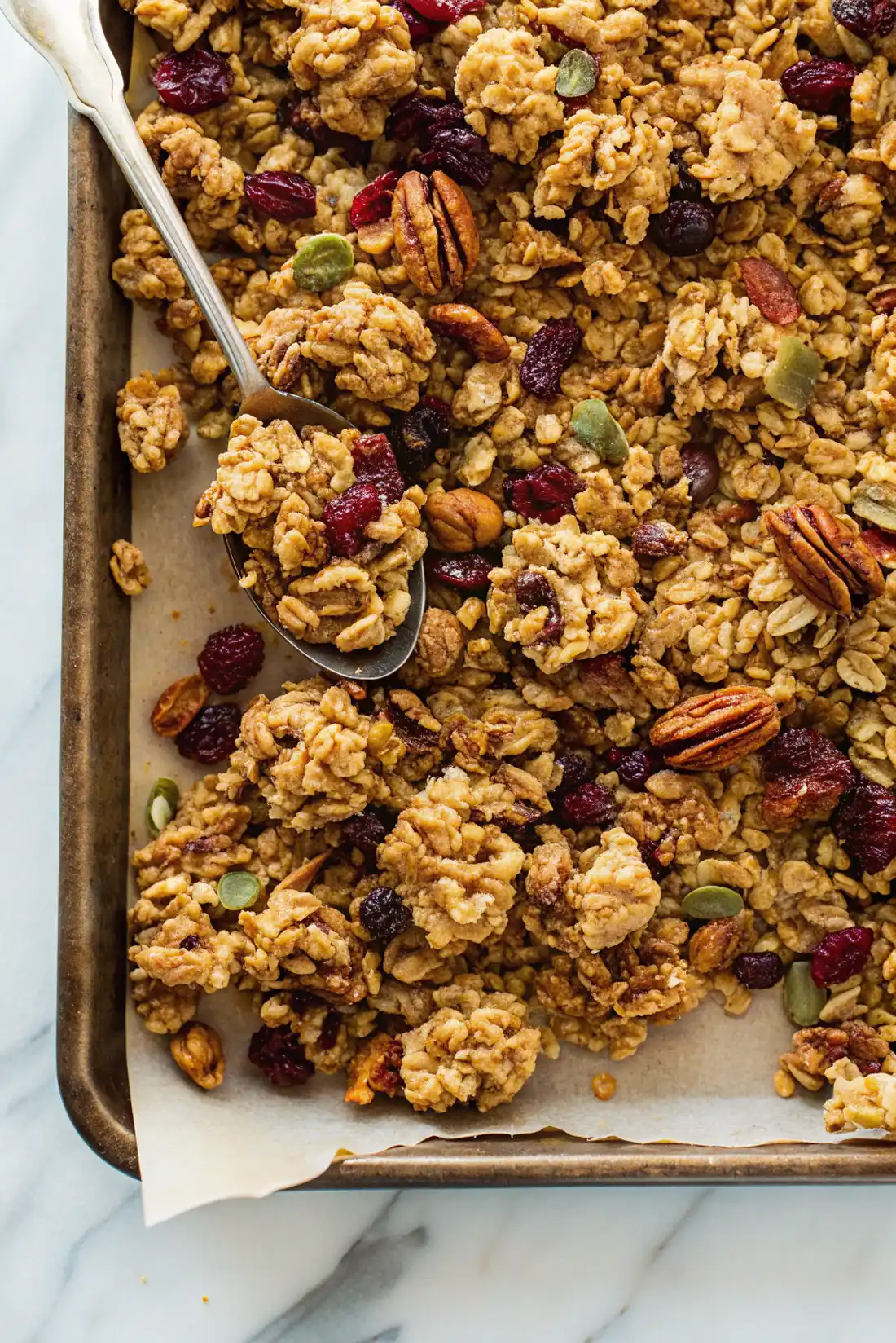 Close-up of clumpy homemade granola with pecans, dried cranberries, and pumpkin seeds on a baking tray with parchment paper.