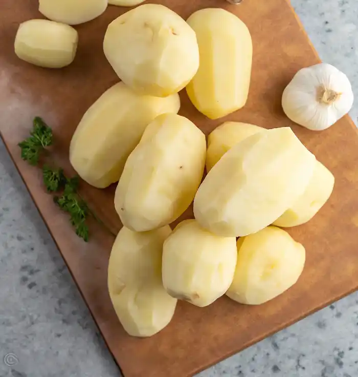 Peeled raw potatoes on a cutting board with fresh garlic and herbs, ready for mashed potatoes