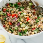 Bowl of chickpea quinoa salad with cucumber, red bell pepper, red onion, and parsley on a marble counter.