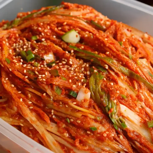 Whole napa cabbage kimchi packed in a plastic container, seasoned with red chili paste, green onions, and sprinkled with sesame seeds.