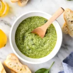 Overhead view of homemade basil pesto in a white bowl with a wooden spoon, surrounded by sliced baguette, fresh basil leaves, lemon, and olive oil on marble surface.