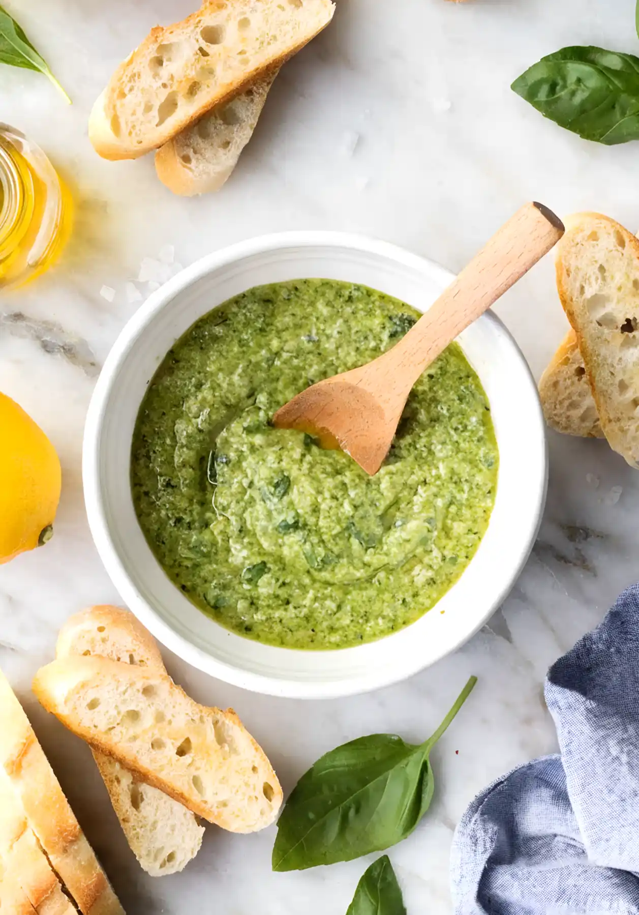 Overhead view of homemade basil pesto in a white bowl with a wooden spoon, surrounded by sliced baguette, fresh basil leaves, lemon, and olive oil on marble surface.