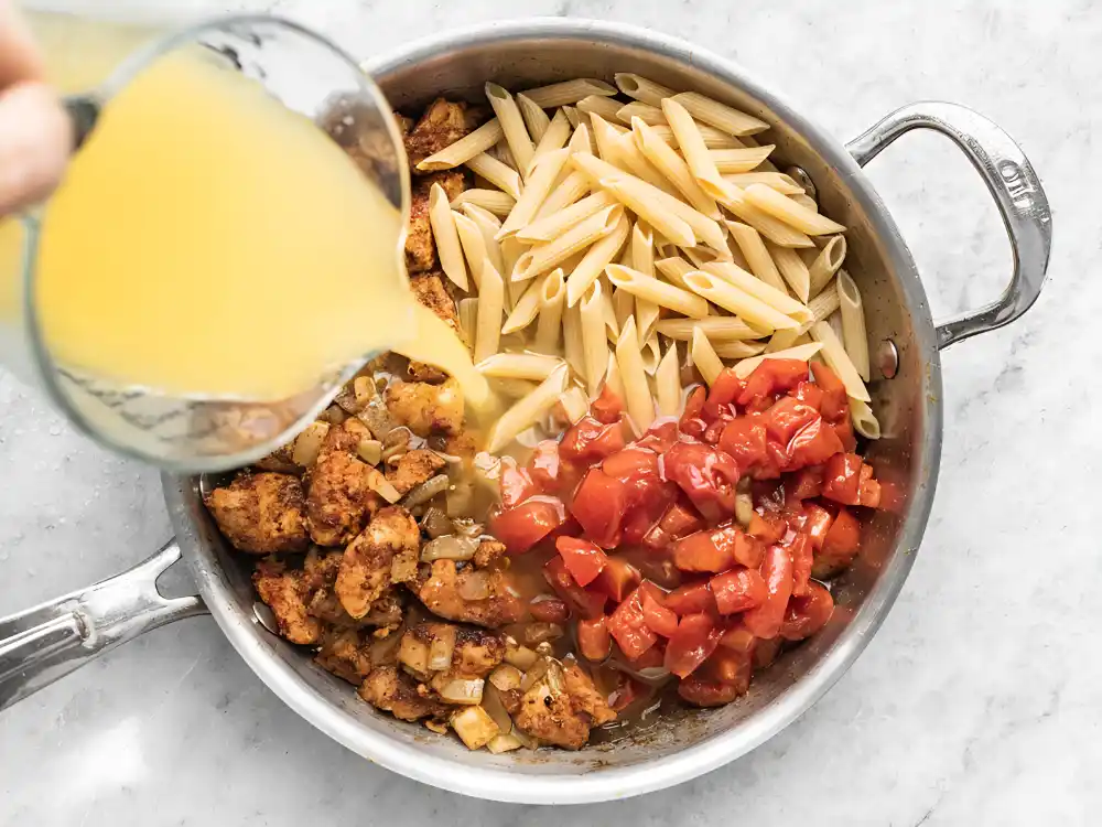 Adding chicken broth to one pot chicken pasta with penne, seared chicken, onions, and fire roasted tomatoes in a skillet.