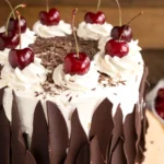 Black Forest cake with whipped cream rosettes, fresh cherries, chocolate shavings, and dark chocolate shards on a rustic wooden stand.