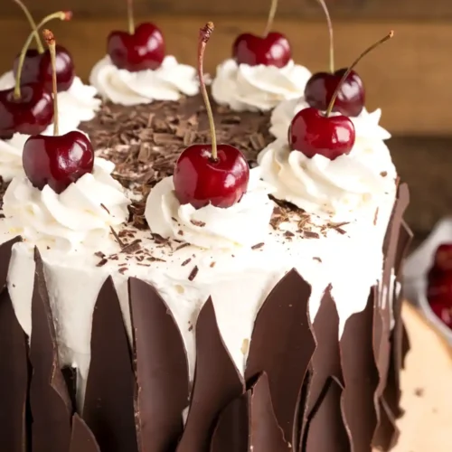 Black Forest cake with whipped cream rosettes, fresh cherries, chocolate shavings, and dark chocolate shards on a rustic wooden stand.