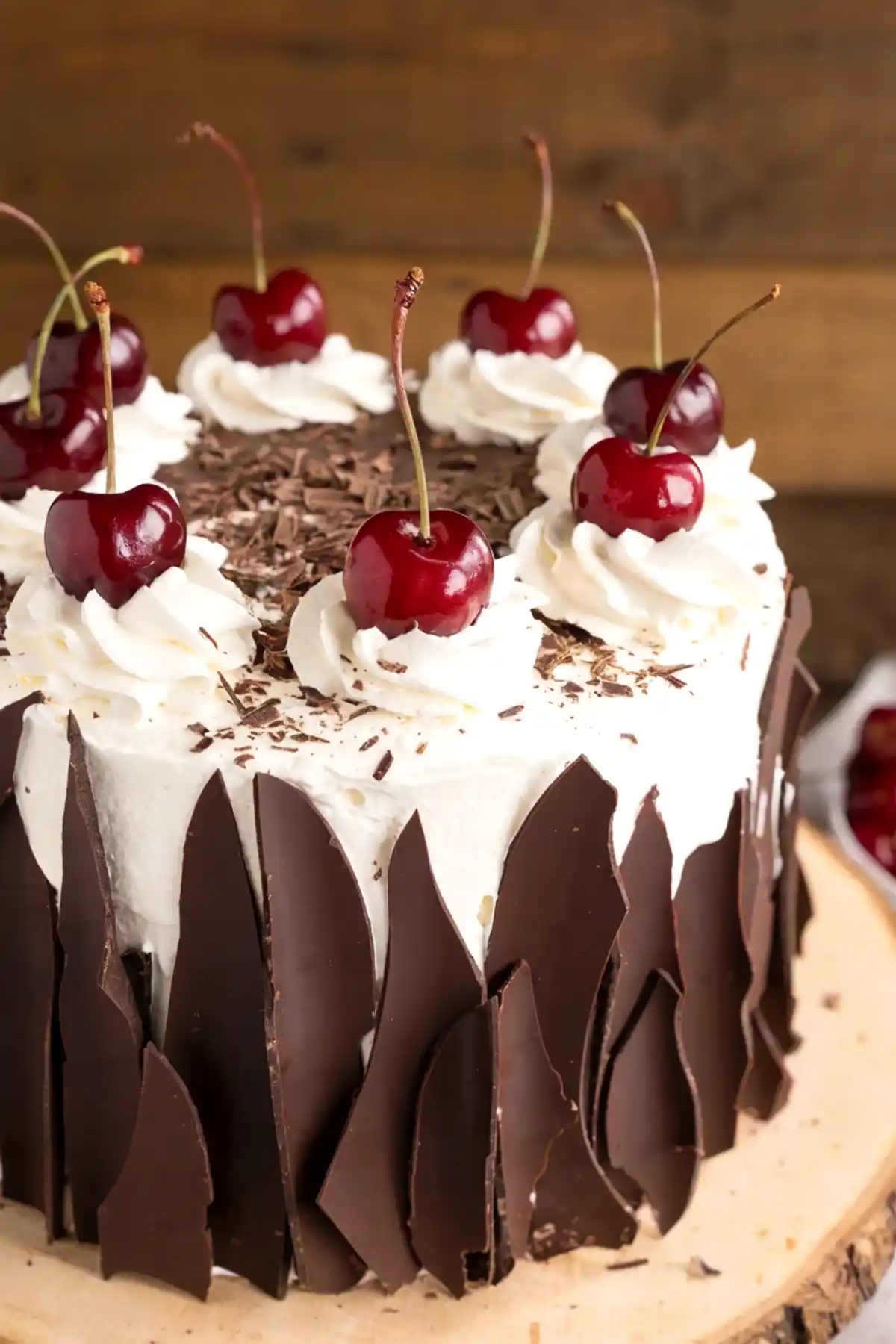 Black Forest cake with whipped cream rosettes, fresh cherries, chocolate shavings, and dark chocolate shards on a rustic wooden stand.