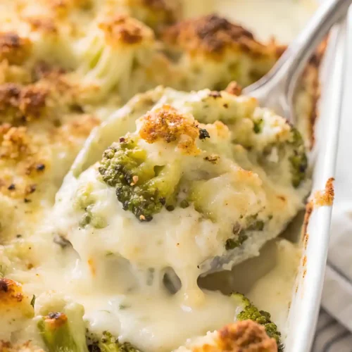Spoon lifting creamy cheesy broccoli casserole with melted white sauce and golden toasted topping in white baking dish.