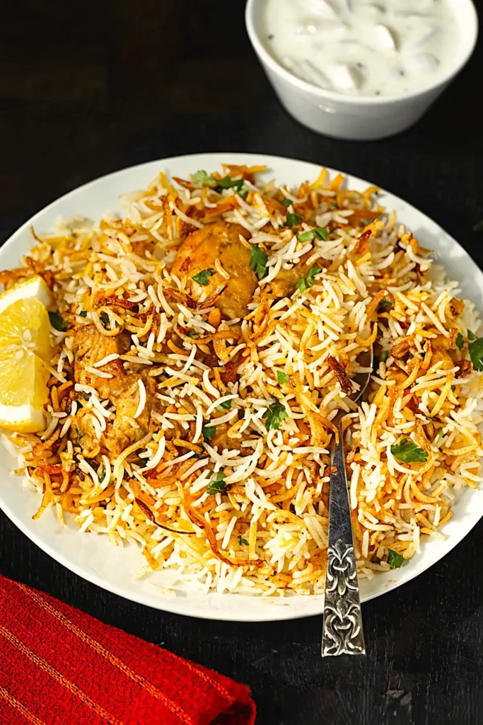 Chicken Biryani Recipe served on a white plate with fluffy basmati rice, crispy fried onions, spiced chicken pieces, lemon wedge, and onion raita.