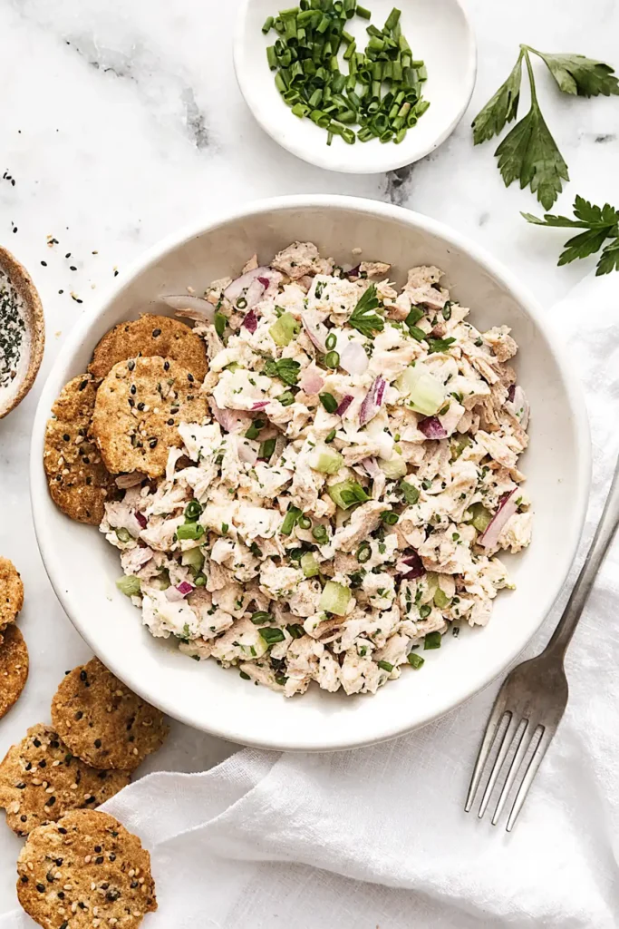 Classic tuna salad in a white bowl with celery, red onion, fresh herbs, and whole grain crackers on the side.