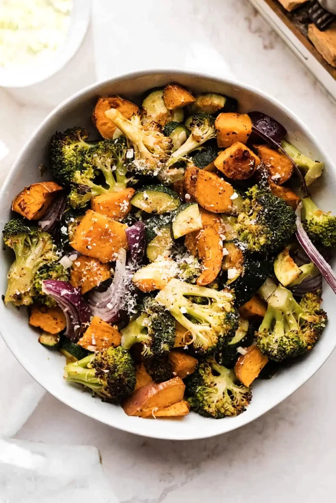 Crispy roasted vegetables in a white bowl with broccoli, sweet potatoes, zucchini, and red onions topped with Parmesan
