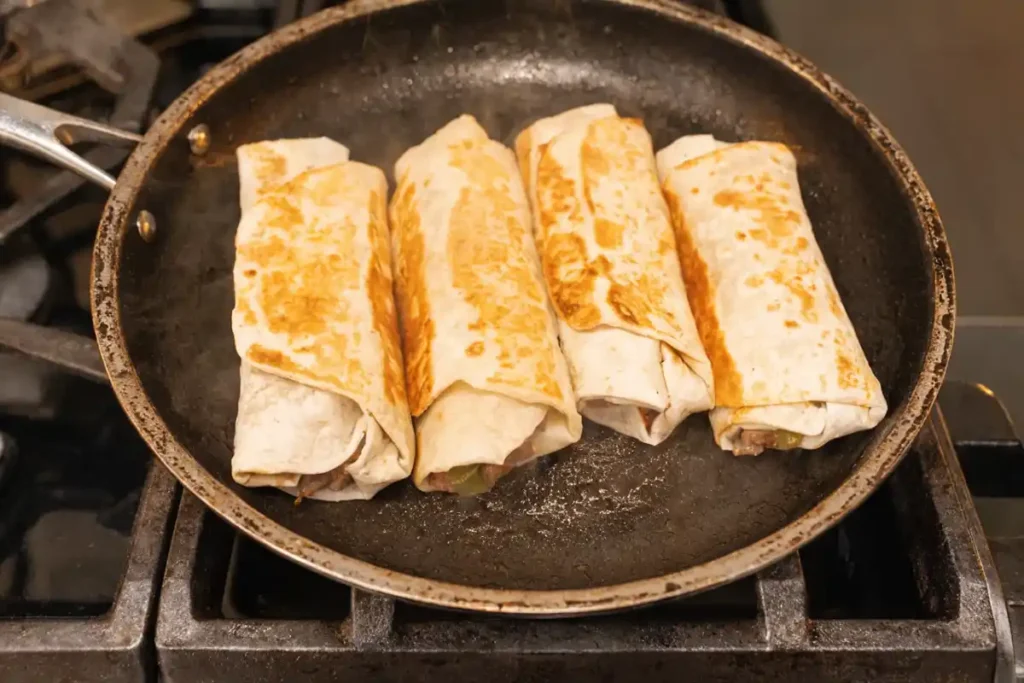 Beef burritos cooking seam side down in a skillet until golden brown and crispy on the stovetop.