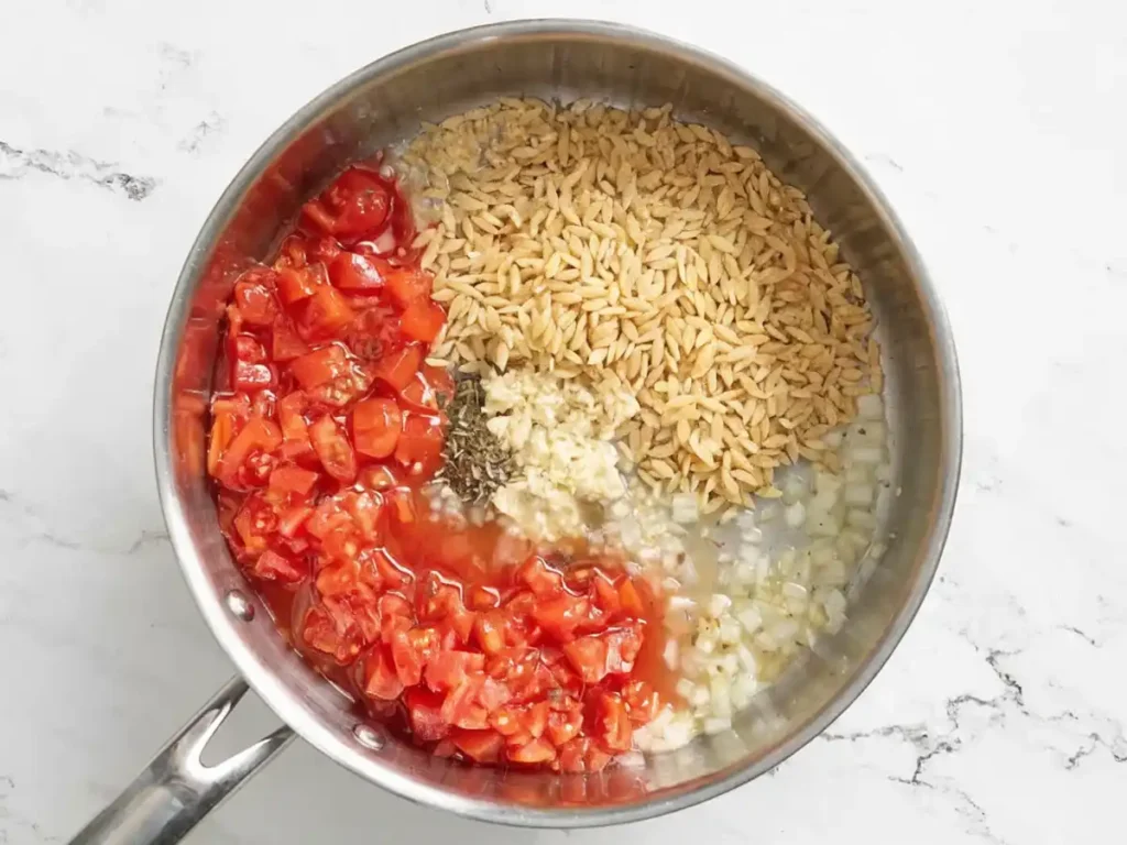 Uncooked orzo with diced tomatoes, chopped onion, garlic, and Italian seasoning in a skillet before cooking.
