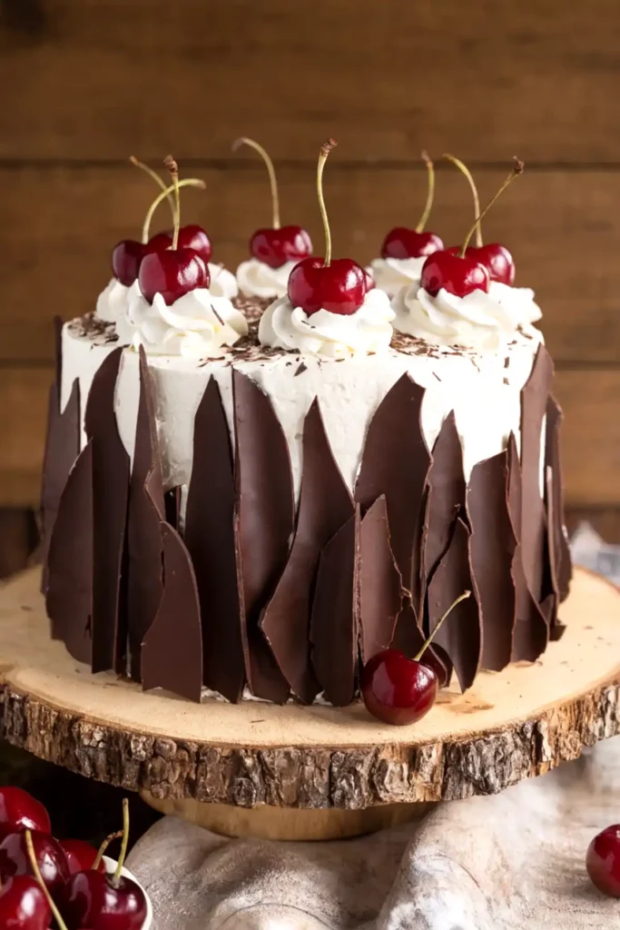 Best Black Forest Cake with whipped cream frosting, fresh cherries, chocolate shavings, and dark chocolate shards on a rustic wooden stand.
