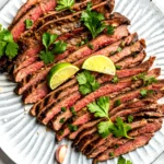 Grilled carne asada sliced against the grain on a ceramic plate with lime wedges and fresh cilantro garnish.