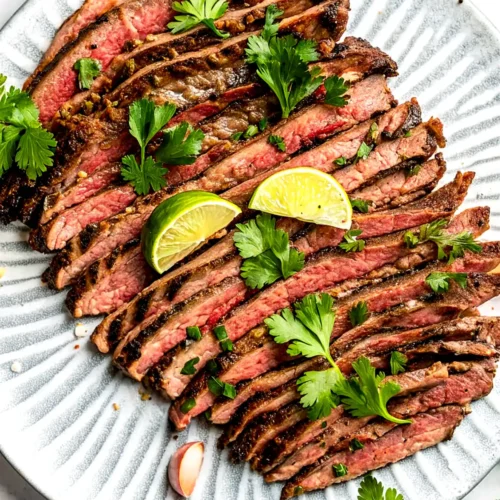 Grilled carne asada sliced against the grain on a ceramic plate with lime wedges and fresh cilantro garnish.