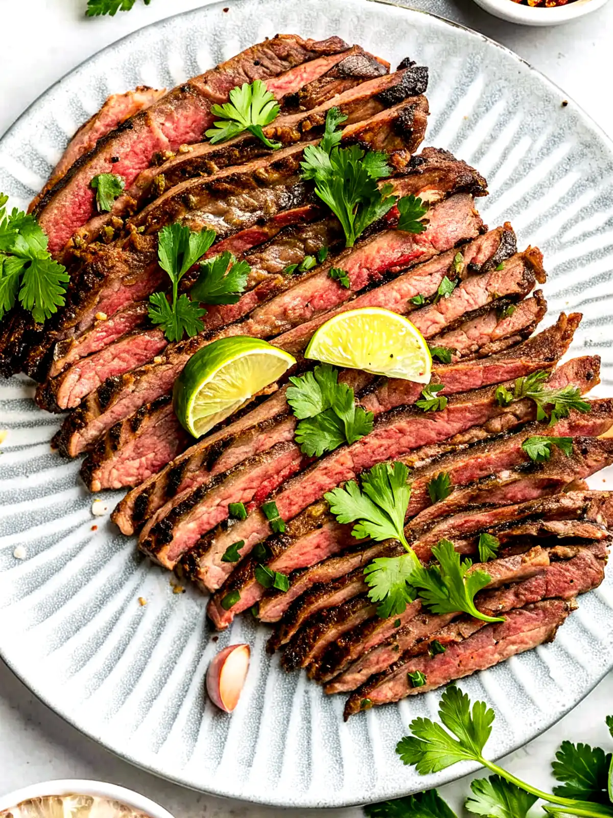 Grilled carne asada sliced against the grain on a ceramic plate with lime wedges and fresh cilantro garnish.