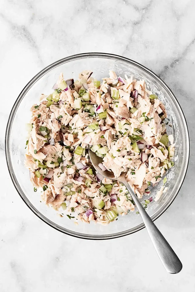 Healthy tuna salad mixed in a glass bowl with celery, red onion, parsley, and light dressing on a marble surface.