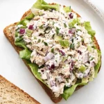Open faced tuna salad sandwich on whole wheat bread with lettuce, red onion, celery, and fresh herbs on a white plate.