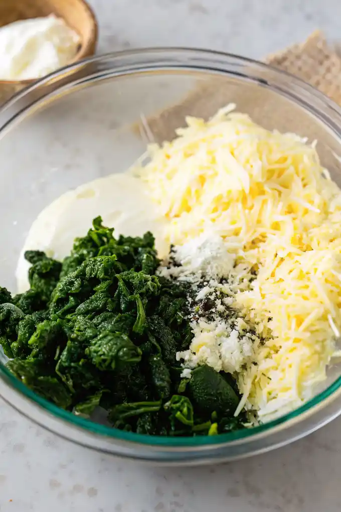 Unmixed ingredients for hot spinach dip in a clear glass bowl with shredded cheese, spinach, and sour cream