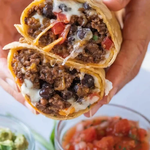 Beef burrito cut in half showing seasoned ground beef, black beans, melted cheese, and diced tomatoes, served with salsa and guacamole.