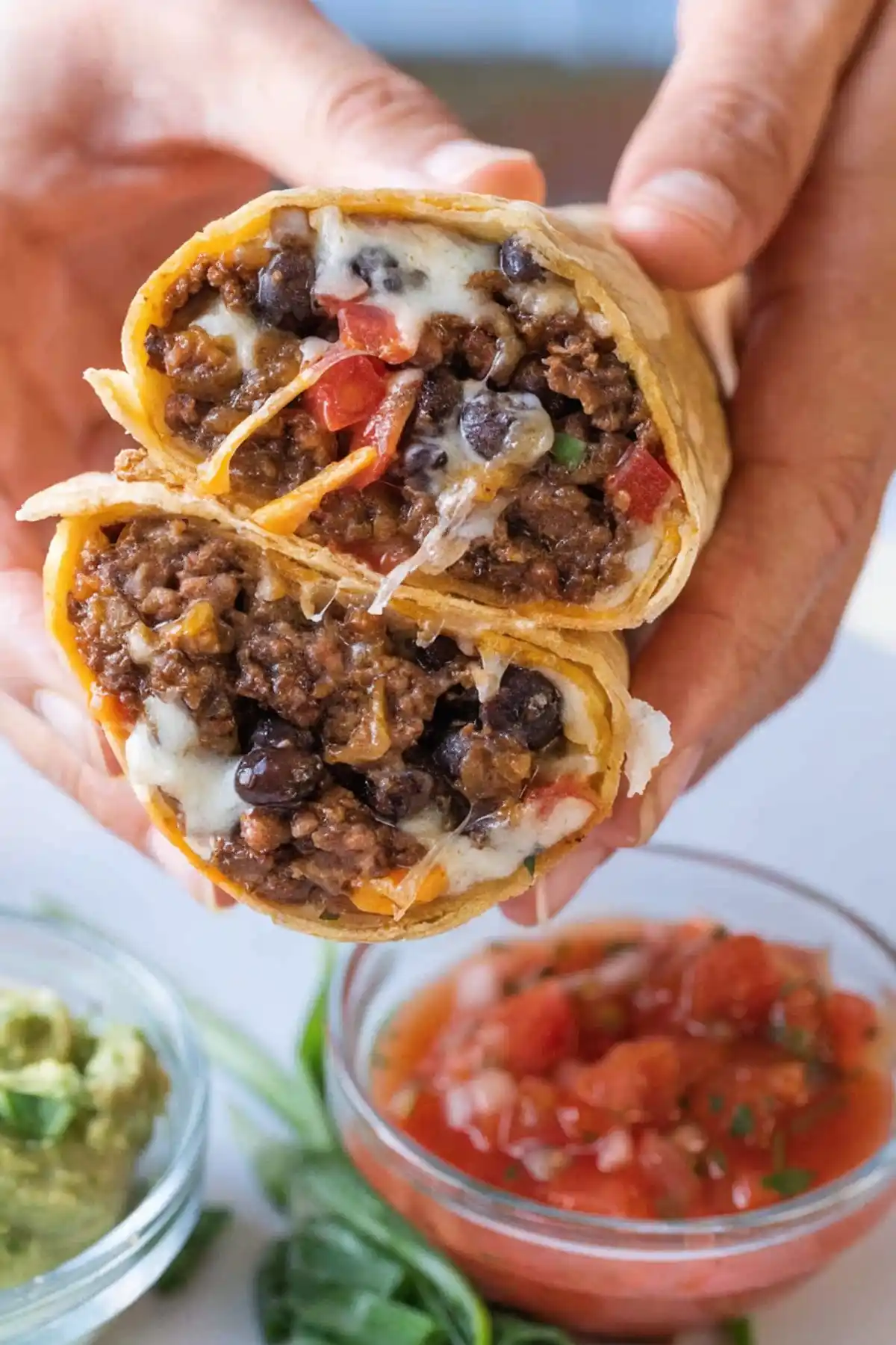 Beef burrito cut in half showing seasoned ground beef, black beans, melted cheese, and diced tomatoes, served with salsa and guacamole.