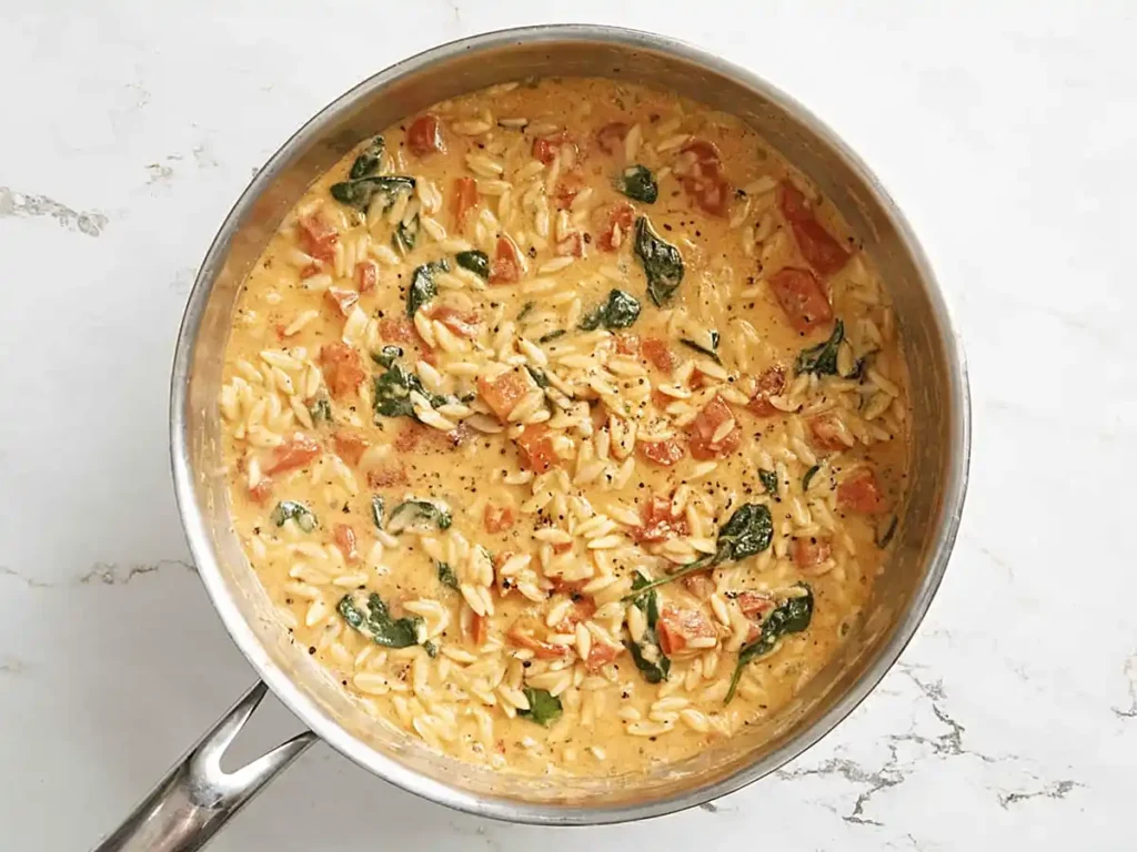 Creamy one pot orzo with spinach and roasted tomatoes cooking in a stainless steel skillet.