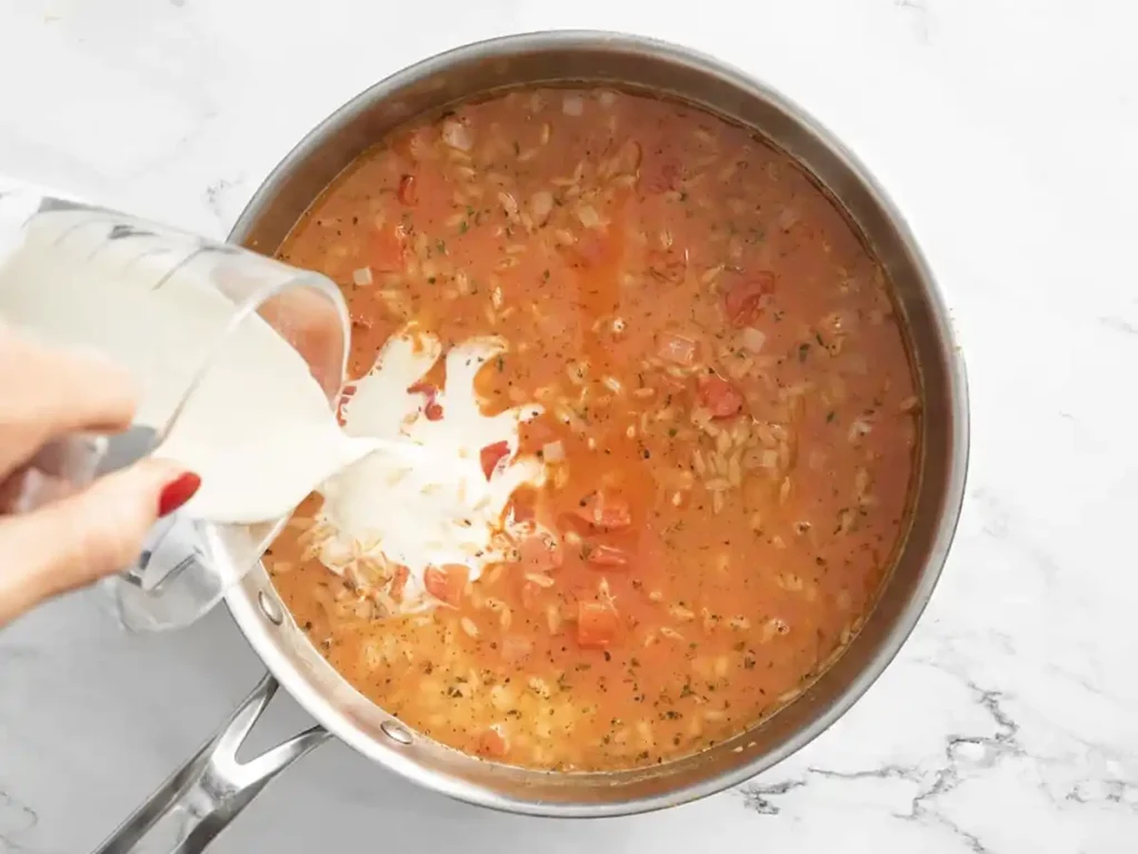 Pouring heavy cream into simmering tomato and orzo mixture in a skillet for creamy orzo recipe.