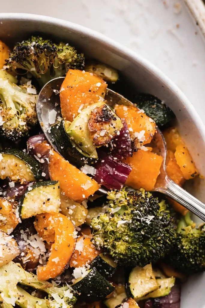 Close up of oven roasted vegetables in a bowl with broccoli, sweet potatoes, zucchini, red onions, and grated Parmesan