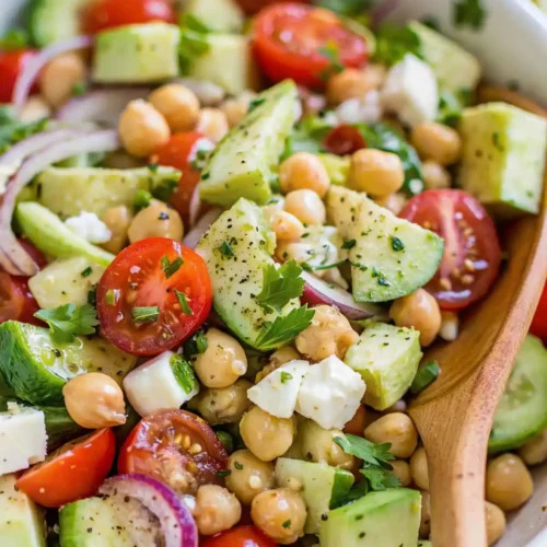 Bowl of Mediterranean chickpea salad with cherry tomatoes, avocado, cucumber, feta cheese, and red onion, tossed with herbs and lemon dressing.