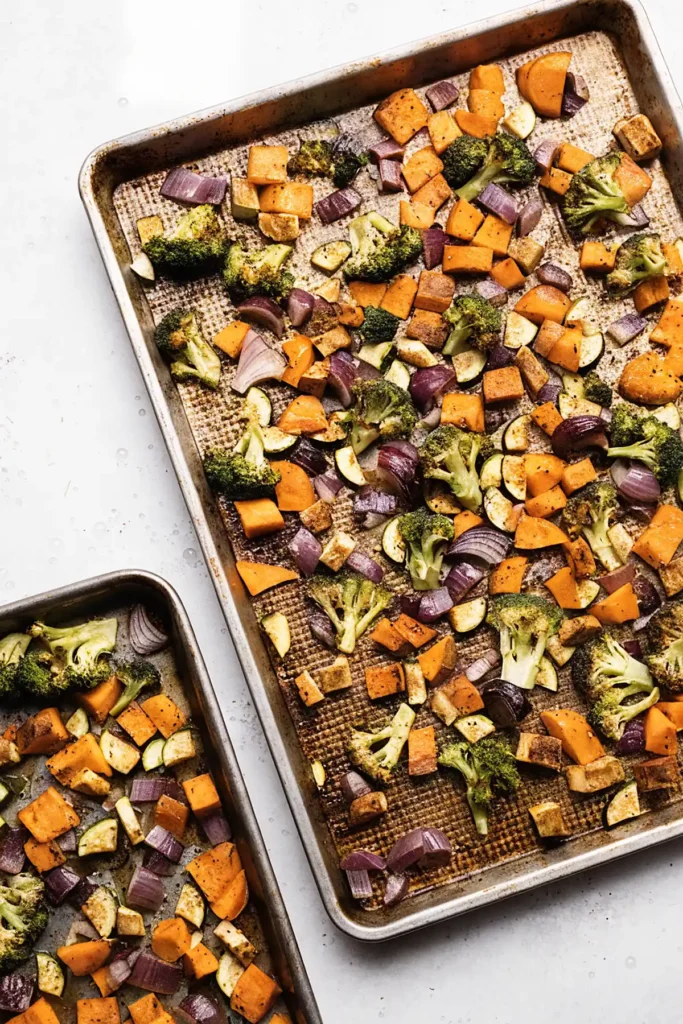 Overhead view of roasted vegetables on sheet pans with broccoli, sweet potatoes, zucchini, and red onions spread in a single layer
