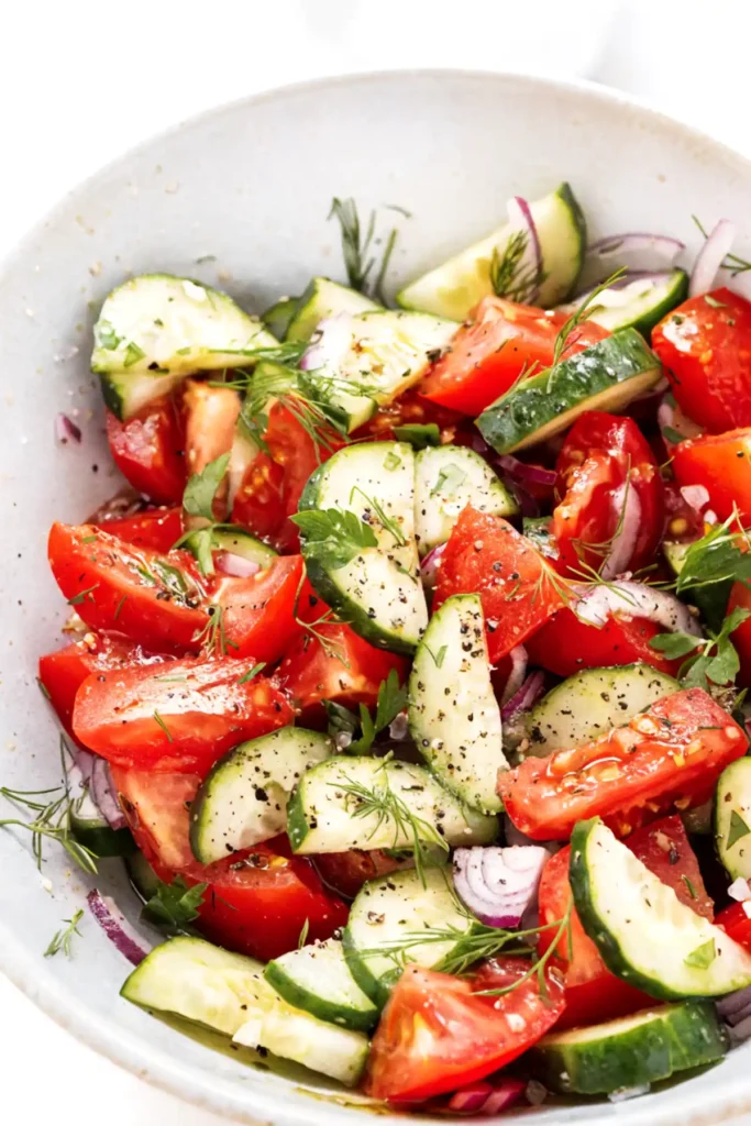 Tomato cucumber salad with fresh dill, parsley, red onion, olive oil, and cracked black pepper in a white bowl.