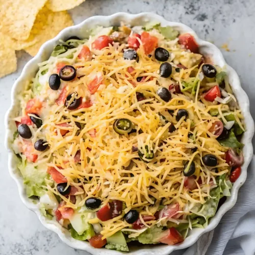 Creamy taco dip in a white scalloped dish topped with shredded cheddar, black olives, diced tomatoes, lettuce, and jalapenos, served with tortilla chips.