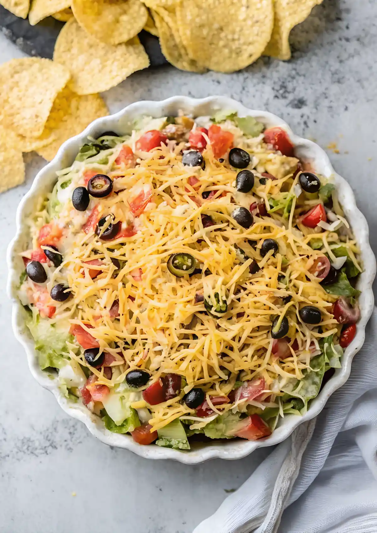 Creamy taco dip in a white scalloped dish topped with shredded cheddar, black olives, diced tomatoes, lettuce, and jalapenos, served with tortilla chips.