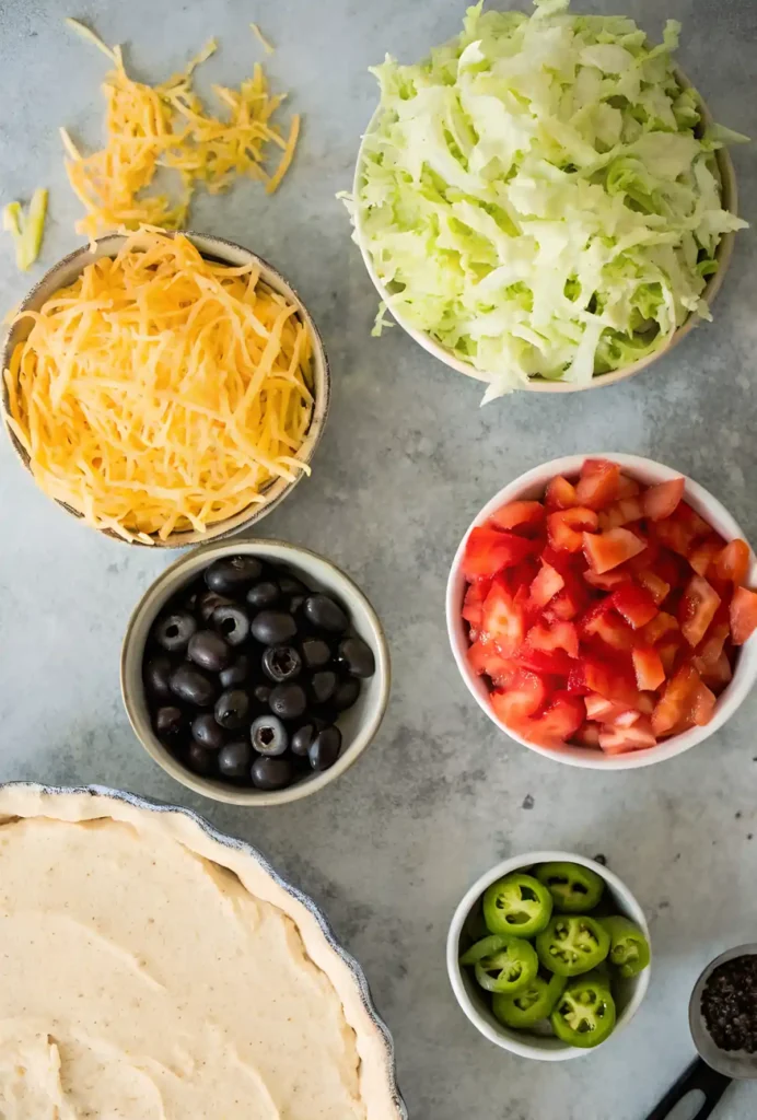 Overhead view of taco dip ingredients including shredded cheese, chopped lettuce, diced tomatoes, black olives, jalapenos, and creamy base in a pie dish.